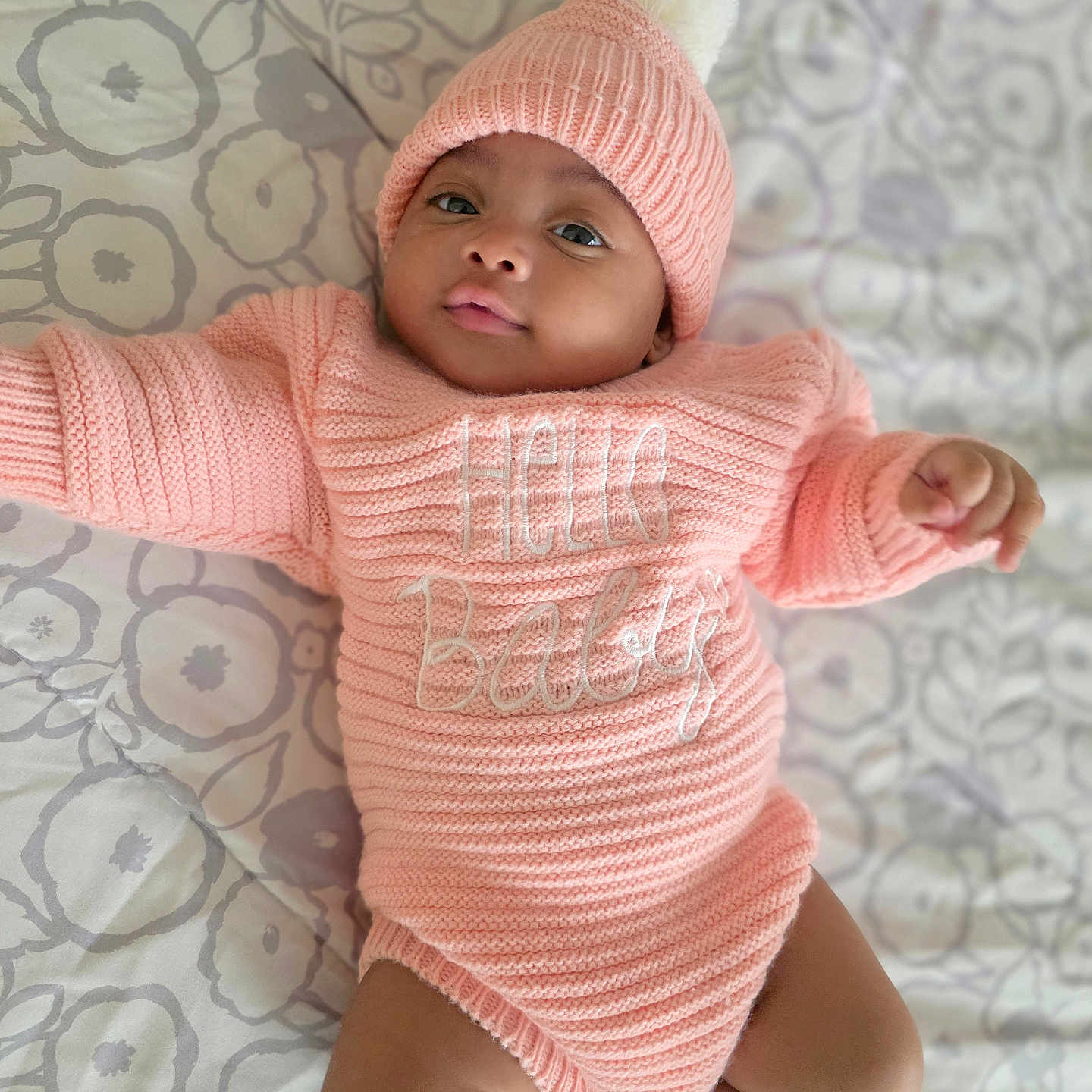 Stacy joined the competition — help win amazing prizes! baby, bedspread, child, clothing, cute, expression, face, floral_pattern, hat, indoors, infant, knitted_outfit, lying_down, pink_knit, pom_pom, portrait, relaxed, skin, soft_lighting, white_socks