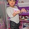 child, toddler, portrait, standing, indoors, playroom, toy_storage, toys, stuffed_animals, doll, purple, pink, hair_bows, gray_sweater, black_pants, socks, smartphone, focused, bedroom, rug