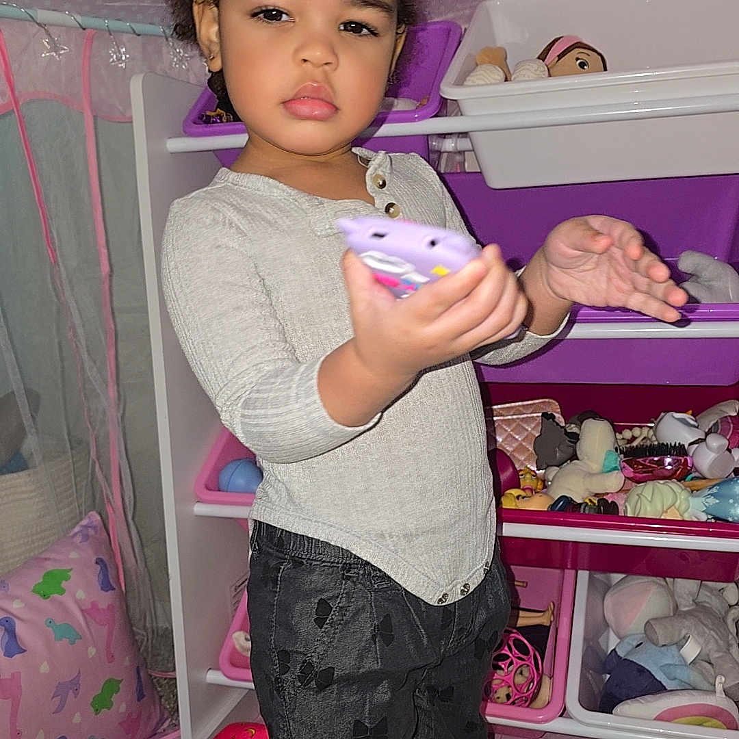 Sᴀᴇʟʟᴇᴍ joined the competition — help win amazing prizes! child, toddler, portrait, standing, indoors, playroom, toy_storage, toys, stuffed_animals, doll, purple, pink, hair_bows, gray_sweater, black_pants, socks, smartphone, focused, bedroom, rug