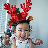 Ridhaan joined the competition — help win amazing prizes! baby, child, reindeer_antlers, christmas_tree, holiday, festive, smiling, happy, hands, white_shirt, indoors, decoration, celebration, red, sparkly, person, cute, face, joyful, holiday_season