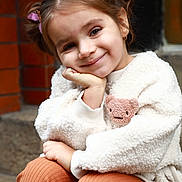 Sofia joined the competition — help win amazing prizes! child, girl, smile, face, hair, butterfly_hair_clip, jacket, teddy_bear_patch, pants, ribbed_pants, sitting, hand_on_face, cute, cozy, outdoor, brick_wall, stone_step, portrait, happy, casual_clothing