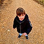 adventure, black_jacket, boy, casual_clothing, casual_shoes, child, curious, daylight, dirt_path, grass, looking_up, nature, outdoor, person, playful, portrait, smile, standing, walking, young
