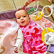 Maelys a rejoint le concours — aidez-le/la à gagner de superbes lots ! baby, child, clothing, colorful, cute, face, floor_tile, hand, happy, indoor, infant, lying_down, person, pink_dress, play_mat, playtime, polka_dot, smiling, soft_toys, toy