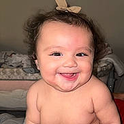Camila joined the competition — help win amazing prizes! baby, smiling, curly_hair, bow, cheeks, diaper, skin, happy, cute, infant, child, indoor, bed, blanket, bassinet, person, face, portrait, chubby, sitting