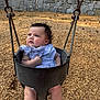 baby, child, swing, playground, wood_chips, bucket_swing, curly_hair, blue_dress, outdoor, stone_wall, infant, seat, metal_chains, cute, expression, legs, summer, daylight, park, young_child