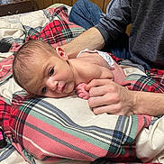 Evalyn joined the competition — help win amazing prizes! baby, infant, hands, blanket, plaid, diaper, bed, wood_paneling, person, skin, comfort, resting, indoor, young_child, support, casual_clothing, blue_jeans, long_sleeve_shirt, closeup, curious