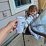 James is registered to the contest to win money with this photo: toddler, child, pacifier, baby_bottle, hand, mug, chair, outdoor, patio, glass_table, window, house_siding, concrete_floor, clothing, person, baby, young_child, reaching, daylight, casual