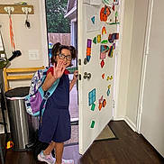 Avelyne is registered to the contest to win money with this photo: child, girl, glasses, backpack, door, colorful_shapes, drawings, wooden_floor, indoor, smiling, waving_hand, sneakers, family_sign, trash_can, chair, key_holder, house, window, light_switch, home