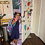 child, girl, glasses, backpack, door, colorful_shapes, drawings, wooden_floor, indoor, smiling, waving_hand, sneakers, family_sign, trash_can, chair, key_holder, house, window, light_switch, home