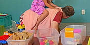 Avelyne joined the competition — help win amazing prizes! child, adult, playing, toys, blocks, wooden_table, pink_dress, indoor, family, fun, crawling, colorful, book, plastic_toys, carpet, container, green_wall, whisk, red_toy, pacifier