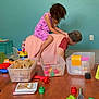 child, adult, playing, toys, blocks, wooden_table, pink_dress, indoor, family, fun, crawling, colorful, book, plastic_toys, carpet, container, green_wall, whisk, red_toy, pacifier