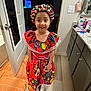 child, smiling, traditional_dress, headpiece, indoor, floor, cabinet, countertop, television, door, light_switch, wall, towel, flooring, person, clothing, happy, portrait, home, young