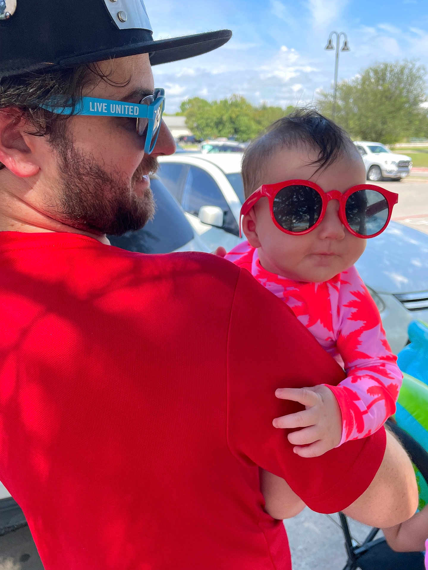 Emilia is registered to the contest to win money with this photo: baby, child, man, sunglasses, hat, outdoor, daytime, car, parking_lot, person, holding, red_clothing, pink_clothing, facial_hair, beard, short_hair, grass, trees, sky, cool
