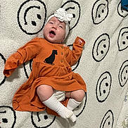 Emilia is registered to the contest to win money with this photo: baby, black, blanket, child, clothing, cozy, cute, flower, happy, headband, indoor, infant, lying_down, orange_dress, portrait, small_person, smiling_face_pattern, soft_texture, white, white_socks