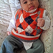 Ezra joined the competition — help win amazing prizes! baby, infant, child, portrait, smiling, tongue_out, sitting, indoor, playful, sweater, orange_clothing, argyle_pattern, pants, chair, cushion, cartoon_bears, nursery, dark_hair, hands, close_up