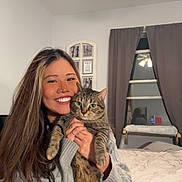 Chairman is registered to the contest to win money with this photo: animal, bed, bedroom, cat, cozy, curtains, face, feline, furniture, happy, holding, home, indoor, long_hair, person, pet, smile, sweater, tabby_cat, window