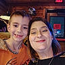 child, adult, selfie, portrait, smiling, happy, indoors, restaurant, wood_paneling, exit_sign, sports_helmet, orange_shirt, black_shirt, necklace, floral_pattern, seated, closeup, faces, family, photograph
