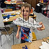 child, boy, smiling, portrait, classroom, school, craft, science_project, foil, aluminum_foil, handmade, creative, art_project, table, scissors, chairs, shelves, classroom_supplies, indoor, standing