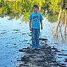 child, boy, smiling, outdoors, pond, water, trees, forest, nature, reflection, lily_pads, mud, wetland, sunlight, blue_shirt, jeans, sneakers, standing, portrait, adventure