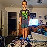 child, boy, smiling, standing, indoors, bedroom, hulk_shirt, superhero, television, dresser, blankets, pillows, backpacks, cat, door, ceiling_fan, furniture, portrait, home, cozy