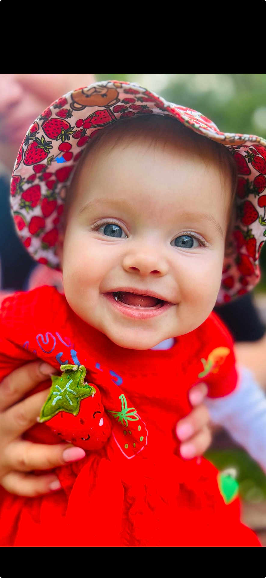 Evelyn is registered to the contest to win money with this photo: baby, child, smiling, blue_eyes, hat, strawberry_pattern, red_dress, cute, portrait, outdoor, person, happy, infant, holding_hands, apparel, closeup, face, headwear, fashion, colorful