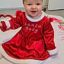 baby, child, smile, red_dress, santa_baby, heart_toy, blanket, indoor, happy, cute, holiday, festive, sitting, white_socks, floor, face, young_child, person, portrait, cozy