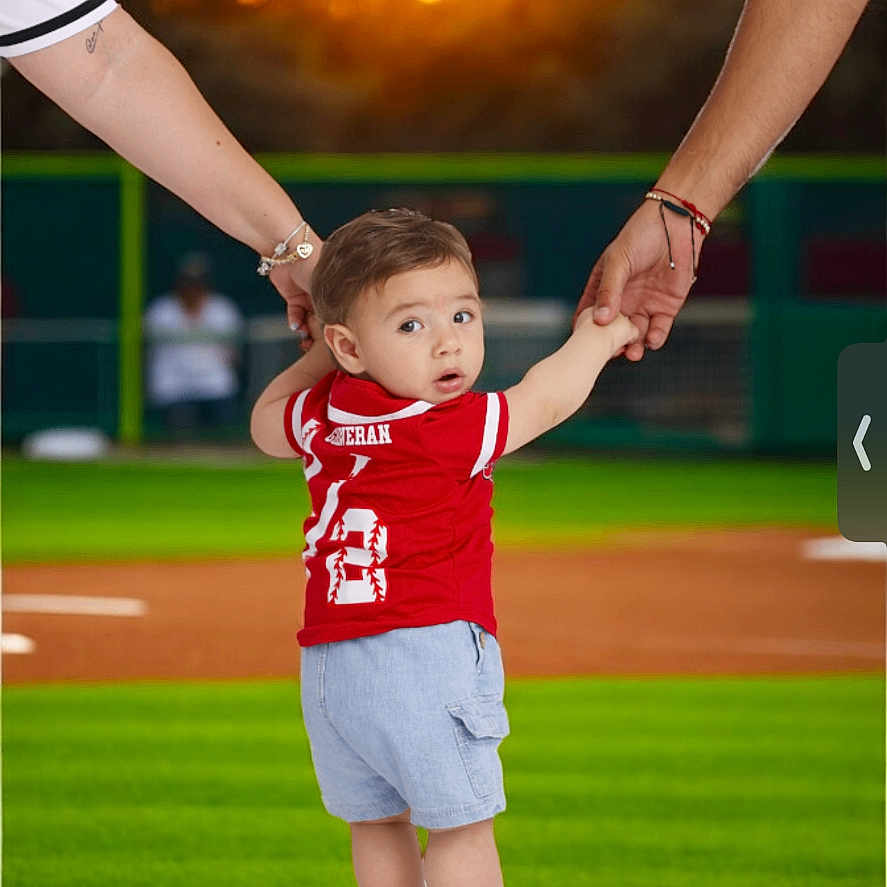Jose is registered to the contest to win money with this photo: adult_arms, athletic_wear, baseball_field, child, curious_expression, dirt, family_moment, green_grass, holding_hands, outdoor, person, recreation, shorts, sneakers, sports_jersey, summer, sunset, team_sport, toddler, young_child