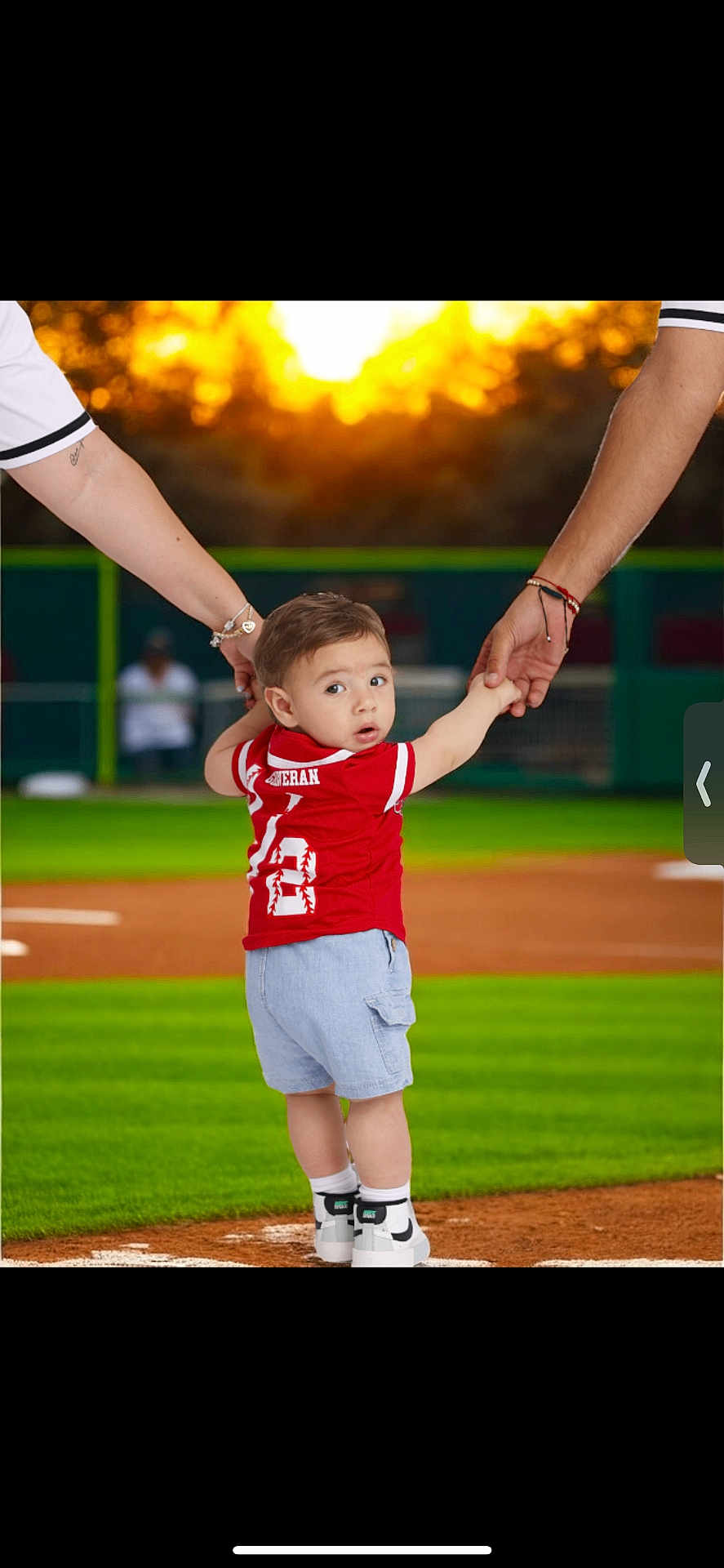 Jose is registered to the contest to win money with this photo: toddler, child, baseball_field, sunset, sports_jersey, shorts, sneakers, holding_hands, adult_arms, green_grass, dirt, outdoor, person, young_child, curious_expression, summer, recreation, team_sport, family_moment, athletic_wear