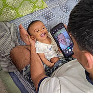 Anvay is registered to the contest to win money with this photo: baby, smiling, person, adult, smartphone, bed, pillow, blanket, indoor, happy, child, onesie, pattern, holding, sitting, carpet, male, shorts, camera, family