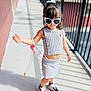 Leah is registered to the contest to win money with this photo: child, toddler, sunglasses, striped_clothing, crop_top, skirt, sneakers, bracelet, sidewalk, sunlight, shadow, fence, urban, fashion, outdoor, person, cute, hair_bow, walking, playful