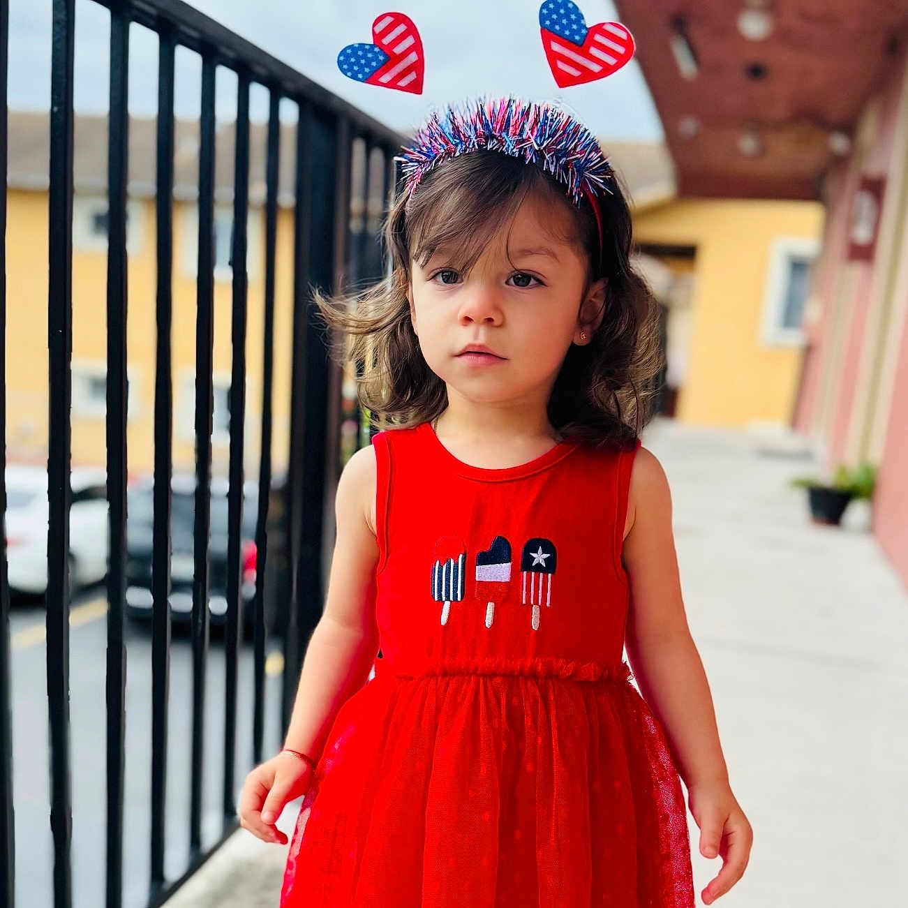 Leah is registered to the contest to win money with this photo: american_flag, balcony, building, child, curious_expression, cute, daylight, girl, headband, heart_shaped_headband, outdoor, patriotic, person, railing, red_dress, short_hair, sidewalk, toddler, urban, window