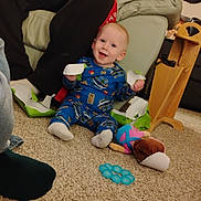 Dominic joined the competition — help win amazing prizes! baby, child, person, pajamas, carpet, toy, lotion_bottle, couch, red_hoodie, living_room, package, tearing, smiling, floor, furniture, indoor, playful, happy, casual, home