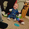 baby, child, person, pajamas, carpet, toy, lotion_bottle, couch, red_hoodie, living_room, package, tearing, smiling, floor, furniture, indoor, playful, happy, casual, home