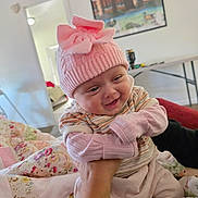 Marley is registered to the contest to win money with this photo: baby, pink_hat, smiling, indoor, quilt, hand, child, cute, knitwear, person, cozy, blanket, apparel, portrait, happy, infant, face, warm, sitting, home