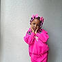 child, girl, pink_clothing, bows, beads, posing, indoor, wall, texture, cute, portrait, fashion, smiling, young, person, standing, colorful, hair_accessories, sweatshirt, pants