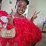 child, girl, red_dress, smiling, happy, pose, hand_gesture, purse, indoor, chair, plastic_chair, hair_puffs, fashion, portrait, seated, carpet, jewelry, necklace, cute, playful