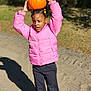 child, pumpkin, pink_jacket, outdoor, sunlight, shadow, sand, path, greenery, fall, autumn, hair_beads, casual_clothing, standing, playful, young_girl, nature, daytime, balance, portrait