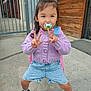 backpack, blue_shoes, casual_clothing, child, concrete, cute, daylight, denim_shorts, gate, outdoor, pacifier, peace_sign, playful, portrait, purple_sweater, smiling, standing, toddler, urban, wooden_wall