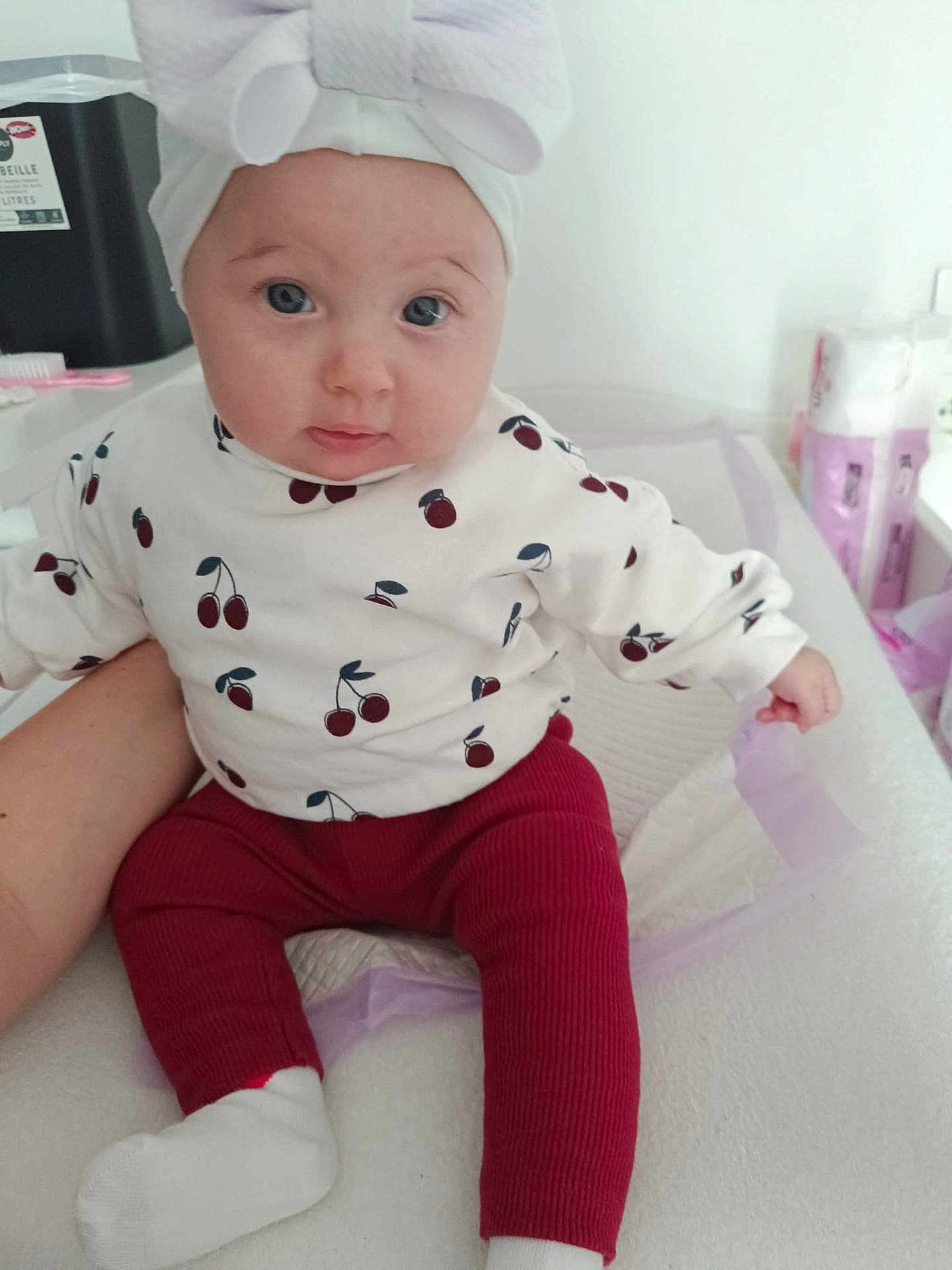 Kayleen a rejoint le concours — aidez-le/la à gagner de superbes lots ! baby, child, headband, bow, cherry_pattern, red_pants, white_socks, infant, cute, portrait, indoor, soft_lighting, supporting_arm, baby_supplies, changing_table, expression, sitting, clothing, person, face