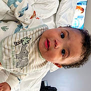 Miguel is registered to the contest to win money with this photo: baby, bed, bib, blanket, child, clothing, curly_hair, cute, elephant, expression, face, fan, home, indoor, pajamas, person, room, television, white_bed, window