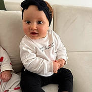 Lya participe au concours pour gagner de l'argent avec cette photo : baby, infant, headband, bow, couch, sofa, white_sweater, black_pants, socks, sitting, big_eyes, chubby_cheeks, hand, portrait, cute, smile, indoors, living_room, neutral_background, soft_light