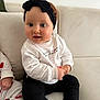 baby, infant, headband, bow, couch, sofa, white_sweater, black_pants, socks, sitting, big_eyes, chubby_cheeks, hand, portrait, cute, smile, indoors, living_room, neutral_background, soft_light