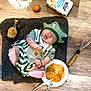 baby, infant, chef_hat, striped_clothing, pancakes, fork, knife, whisk, egg, maple_syrup, flour, plate, wooden_floor, cooking, food, nap, cute, sleeping, kitchen, recipe
