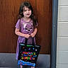 child, girl, purple_dress, striped_clothing, tote_bag, school, summer, purple_sandals, door, wooden_door, concrete_floor, indoor, smiling, standing, casual_clothing, cute, young, person, footwear, holding_bag