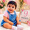 baby, girl, smiling, denim_dress, pink_shirt, white_shoes, flowers, pink_bow, fairy_lights, fluffy_rug, sign, milestone, portrait, cute, indoor, celebration, sitting, happy, headband, decor
