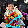 baby, car_seat, toy, child, smile, clothing, jacket, sock, car_interior, seatbelt, headrest, window, cupholder, purple_bag, plastic_toy, child_protection, cute, infant, portrait, happy