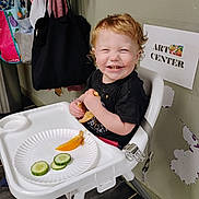 Timothy is registered to the contest to win money with this photo: toddler, high_chair, smiling, bread, cucumber, orange, paper_plate, food, indoor, child, red_hair, curly_hair, black_shirt, wall, bags, hooks, sign, art_center, floor, happy