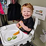 toddler, high_chair, smiling, bread, cucumber, orange, paper_plate, food, indoor, child, red_hair, curly_hair, black_shirt, wall, bags, hooks, sign, art_center, floor, happy