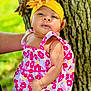 baby, child, flower_headband, yellow_headband, pink_flower_dress, tree, outdoor, nature, green_background, person, infant, cute, portrait, skin, hand, summer, sunlight, colorful_clothing, closeup, baby_fashion