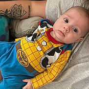 Logan is registered to the contest to win money with this photo: baby, child, person, tattoo, arm, clothing, cowboy_outfit, yellow_shirt, blue_pants, indoor, sofa, couch, casual, portrait, infant, cute, relaxing, human, face, expression